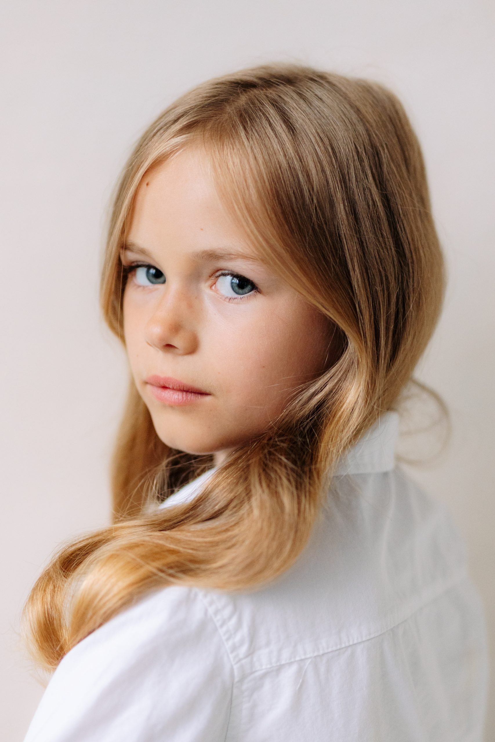 Portrétní fotografie malé dívky s dlouhými světlými vlasy a modrýma očima v bílé košili. Jemné světlo a přirozený výraz zachycený v HLight Studiu fotografem Lukášem Vavřincem.